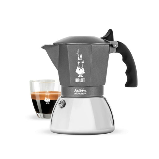 A Bialetti Brikka Induction Coffee Pot with a sleek design, featuring an aluminum top and stainless steel base, shown with a glass of freshly brewed espresso.